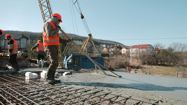 Construction worker in orange vest levels wet concrete slab with long wooden tool, Outdoor building site in residential area with visible crane and workers in background