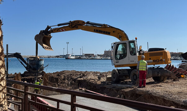 Budva,Montenegro-April 11,2025:Liebherr and Volvo excavators at a waterside construction site with workers and a marina in the background.  

