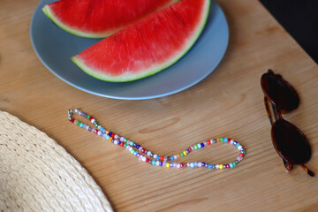 Plate with watermelon slices, glass of orange juice or cocktail, straw hat, colorful beaded necklace and sunglasses on the wooden table. Summer essentials concept. Selective focus.