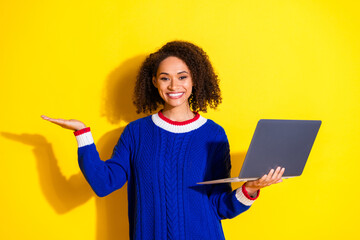 Confident young woman in a blue sweater holding a laptop and gesturing on a vibrant yellow...