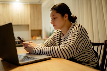 A young woman with a cell phone in hand and a laptop sits at the dining room table in her kitchen...