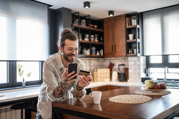 Freelancer smiling and buying online with credit card and smartphone in kitchen