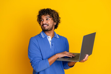 Smiling young man in blue shirt holding laptop against bright yellow background, representing...