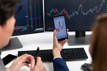 Cropped pointing hand with pen in businessman, checking with holding smartphone by woman investor online website program application with dynamic stock market at home office at back side. Infobahn.