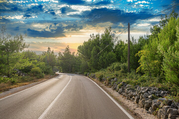 A quiet winding road cuts through a lush green forest under a dramatic sunset sky, evoking a sense of peace and natural beauty.