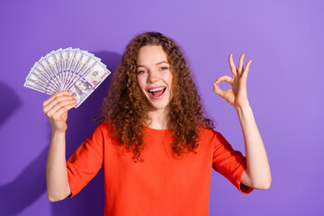 Young cheerful woman with curly hair gestures an okay sign while holding a fan of hundred-dollar...