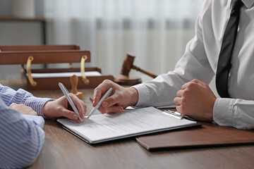 Notary pointing at document and client putting signature at wooden table indoors, closeup