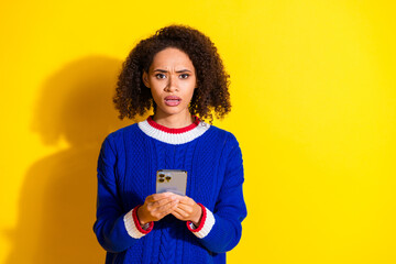 Young woman with curly hair in blue sweater displaying surprise while using smartphone against...