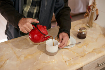 Farmer pouring steaming tea from crimson teapot into ceramic mug, homemade pastry and jam nearby, warm morning light illuminating rustic kitchen counter