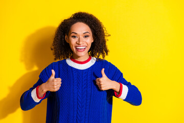 Smiling young woman in a colorful knit sweater gesturing thumbs up against a vibrant yellow...