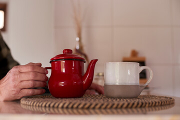 Elderly man gently holding a vintage red teapot, preparing a comforting morning tea ritual, with a ceramic mug placed nearby on a woven mat in a cozy kitchen setting