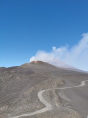 Curved gravel path crosses barren volcanic terrain toward the smoking summit of Mount Etna, with dark slopes under a vivid blue sky in Sicily, Italy