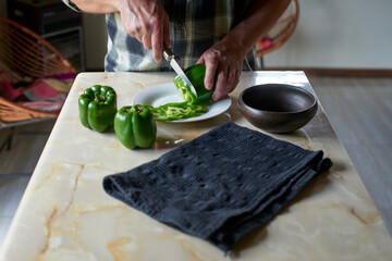 Chef slicing fresh green bell peppers on kitchen counter, dark gray towel and clay bowl nearby, preparing ingredients for cooking