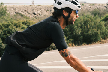 Profile view of a male cyclist in black cycling apparel, wearing a helmet and sunglasses, riding on...