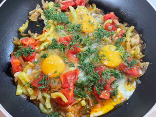Fried eggs in the process of cooking. Three raw eggs broken in a black frying pan on a layer of already fried onions, peppers, tomatoes and sprinkled with fresh dill on top, close-up, top view.