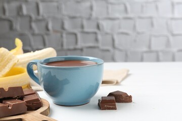 Tasty chocolate milk in cup, pieces and banana on white wooden table, closeup. Space for text