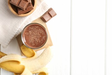 Tasty chocolate milk in glass, banana and pieces on white wooden table, flat lay