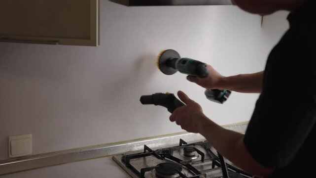 Professional cleaning specialist wiping greasy kitchen wall surface using steam cleaner and rotating drill brush for thorough surface sanitization and stain removal. Concept of housekeeping, cleaning.