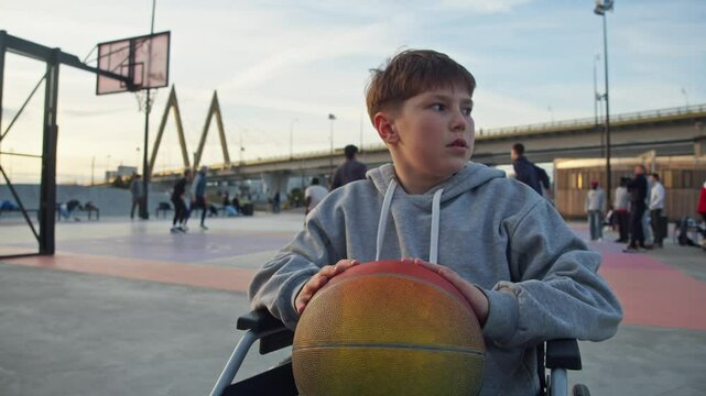 Sad and upset young boy with special needs, sitting in a wheelchair, is seen against the backdrop of his peers playing basketball in an outdoor playground.