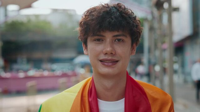 Portrait of happy man wrapped with rainbow LGBTQ flag standing on city street. Smiling young guy expresses proudness of sexual orientation outdoors. Support of minorities community