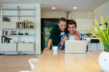 A young married couple is talking to parents, family and friends on a video call via a laptop while sitting in the living room of their modern house.