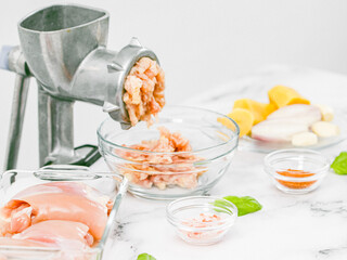 Manual meat grinder with glass bowl,boneless chicken thighs,peeled onions,garlic on a marble table .