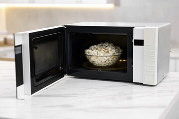 Open microwave oven with bowl of popcorn on white marble table in kitchen