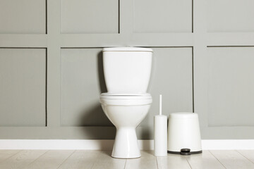 Ceramic toilet bowl, brush and trash can near grey wall indoors