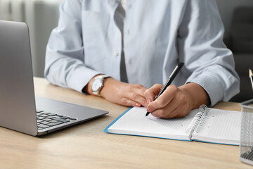 Woman learning online using laptop and taking notes at wooden table indoors, closeup. Self-study
