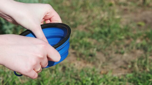 Girl unfolds a collapsible silicone bowl for animals and puts it down. Close-up. Traveling with animals. Walking on a sunny day in a city park. Care concept, animal life
