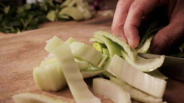 Side view of hands slicing fresh Pak Choi on a wooden board. Close-up food prep in warm kitchen light with leafy greens blurred in the background.