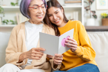 Smiling asian adult daughter greeting happy senior mother with mother's day presenting bouquet of flowers and card,happy elderly mom and grown up child hug embrace celebrate birthday at home together