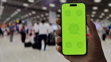 GREEN SCREEN CHROMA KEY A hand holding a smartphone with a green screen in an airport terminal. Blurred travelers and modern architecture in the background