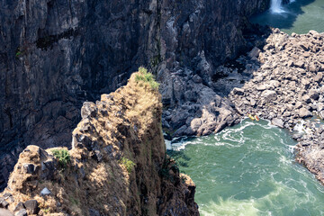 Rocky outpost in the victoria falls canyon with sun shining on it and the green rapids below 