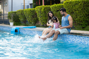 Young indian parents and little child daughter enjoy splashing water at swimming pool in summer holiday, India Family having fun relaxing together at hotel resort vacation. Bonding and togetherness