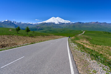 The amazing view to peak of Mount Elbrus, the road, green meadows and Caucasian ridge.