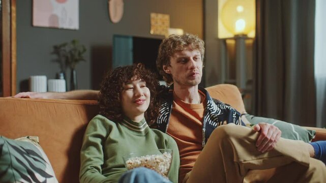 Young loving couple relaxing together on comfortable sofa, watching movie and sharing bowl of popcorn in cozy living room with warm lighting