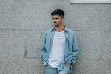 Obraz premium A young man wearing a shirt and jeans standing in front of a wall with his hands in his pockets, presenting a casual and confident appearance.