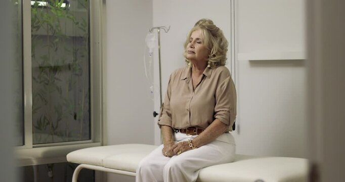 Senior woman sitting in clinic exam room touching neck, appearing concerned while waiting for medical consultation with IV equipment in background