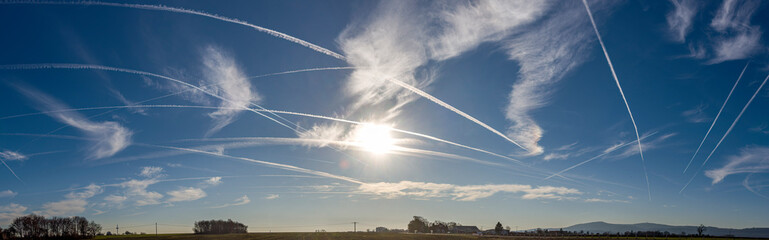 Zarte Wolken mit Kondensstreifen von Flugzeugen bei klarer Luft  vor blauem Himmel, mit Sonne und...