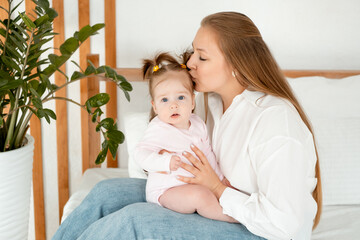 a mother with a baby daughter, a mother gently hugs her baby girl kissing, maternal love and care, a happy family with a baby on the bed at home in the bedroom.