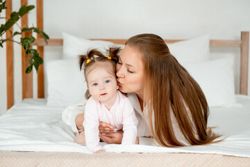 a mother with a baby daughter, a mother gently hugs her baby girl kissing, maternal love and care, a happy family with a baby on the bed at home in the bedroom.