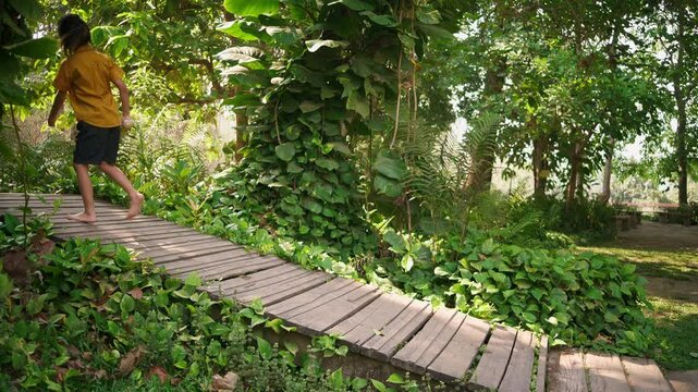 Child walks barefoot from right to left along a slightly sloped wooden path surrounded by lush greenery. Calm and natural outdoor moment.
