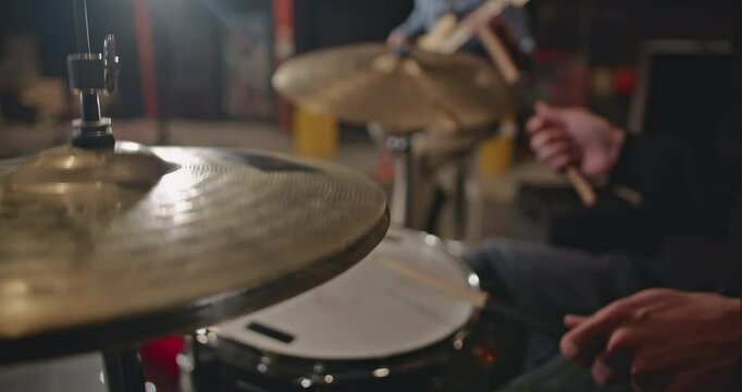 Close-up of a drummer playing the hi-hat and snare drum in a dimly lit room with a guitarist in the background.