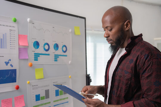 Businessman is analyzing and comparing marketing data displayed on a whiteboard, taking notes on a clipboard and preparing a presentation for a meeting