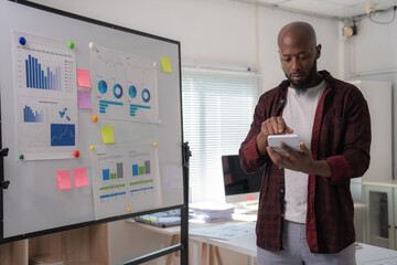 Businessman using a tablet to analyze data in a modern office, with charts and graphs on a whiteboard highlighting business strategy and performance analysis