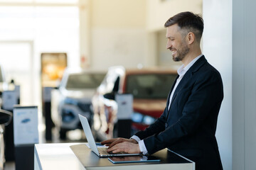 Smiling salesman in formal suit using laptop in bright modern car dealership showroom, handsome...