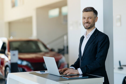 Portrait of smiling Caucasian millennial car salesman working on laptop at car dealership showroom, handsome young man in suit standing at desk with computer and looking at camera, copy space