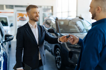 Smiling salesman giving car keys to male customer in bright modern car dealership showroom, auto...
