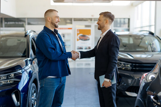 Salesman shaking hands with smiling male customer in a car dealership showroom, greeting with purchase, luxury vehicles displayed around them, happy young man buying new auto